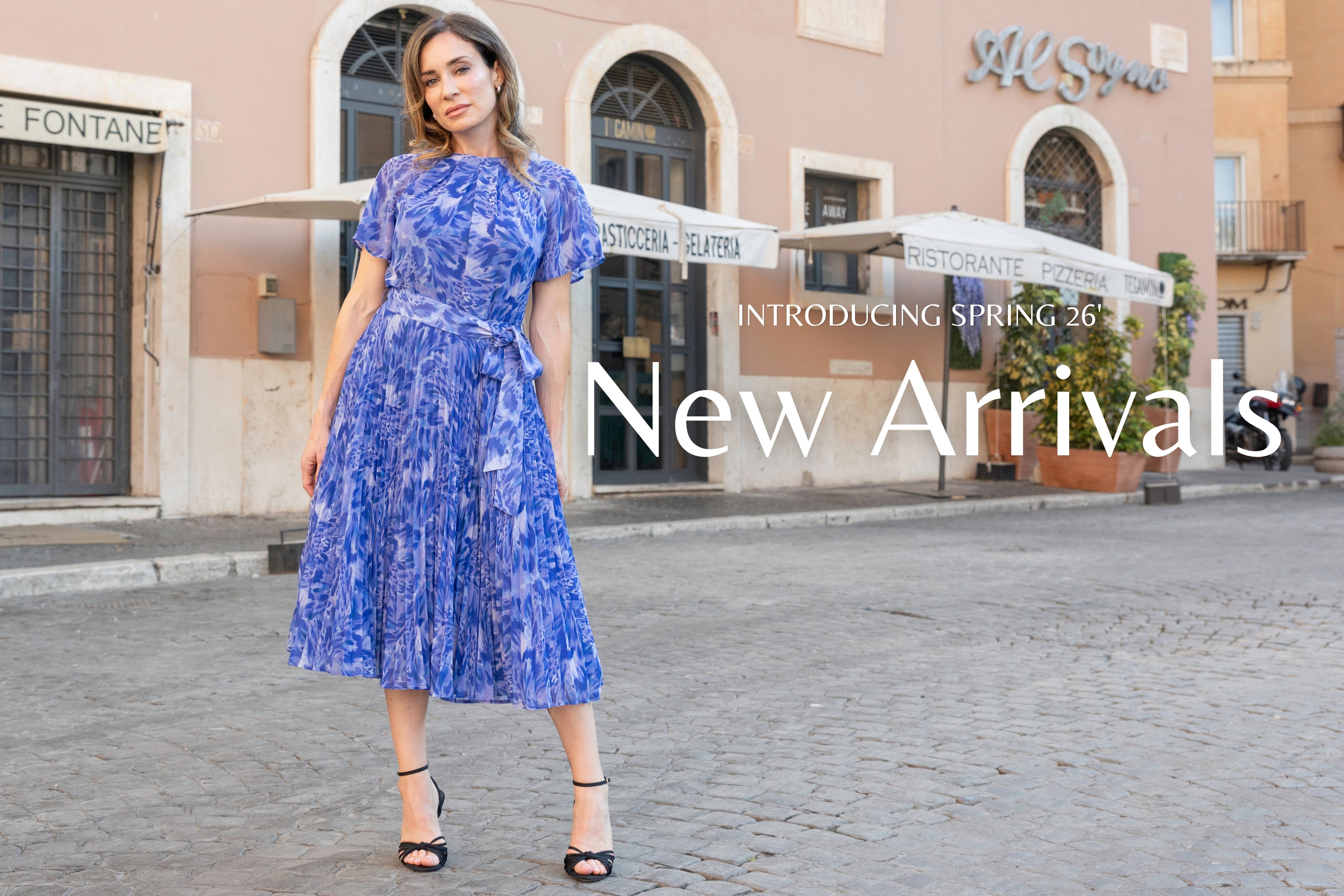 A woman modeling a blue and white patterned midi dress with short puff sleeves and a tie waist, standing on a cobblestone street in front of a restaurant.