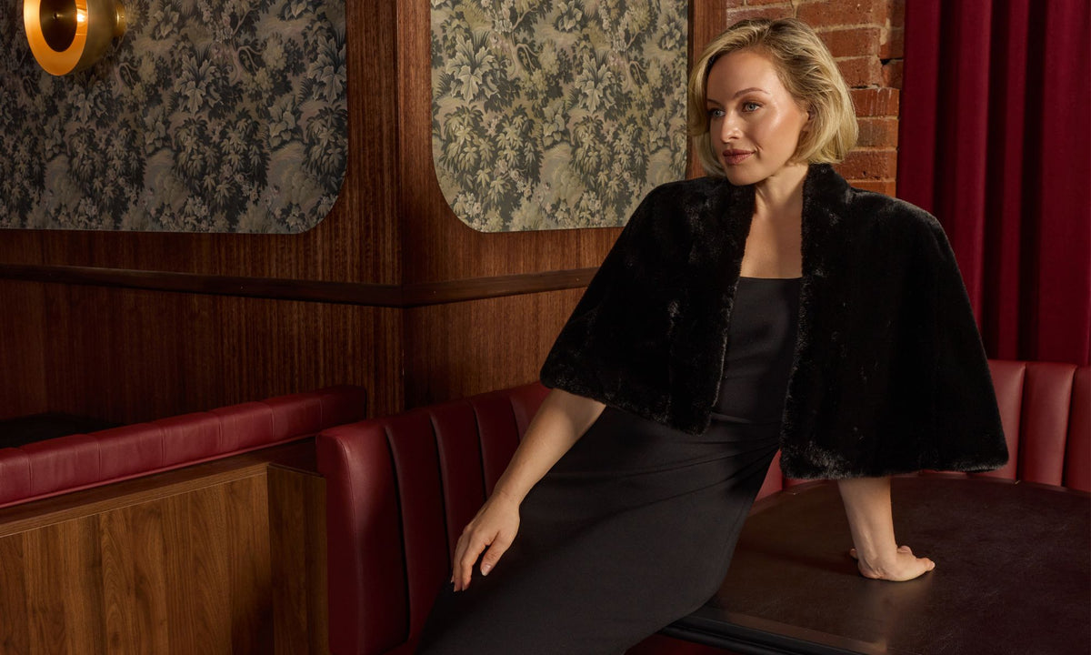 Woman sitting on a red booth in a bar with patterned walls and a brick wall. Avila Black Gown & Gia Black Faux Fur